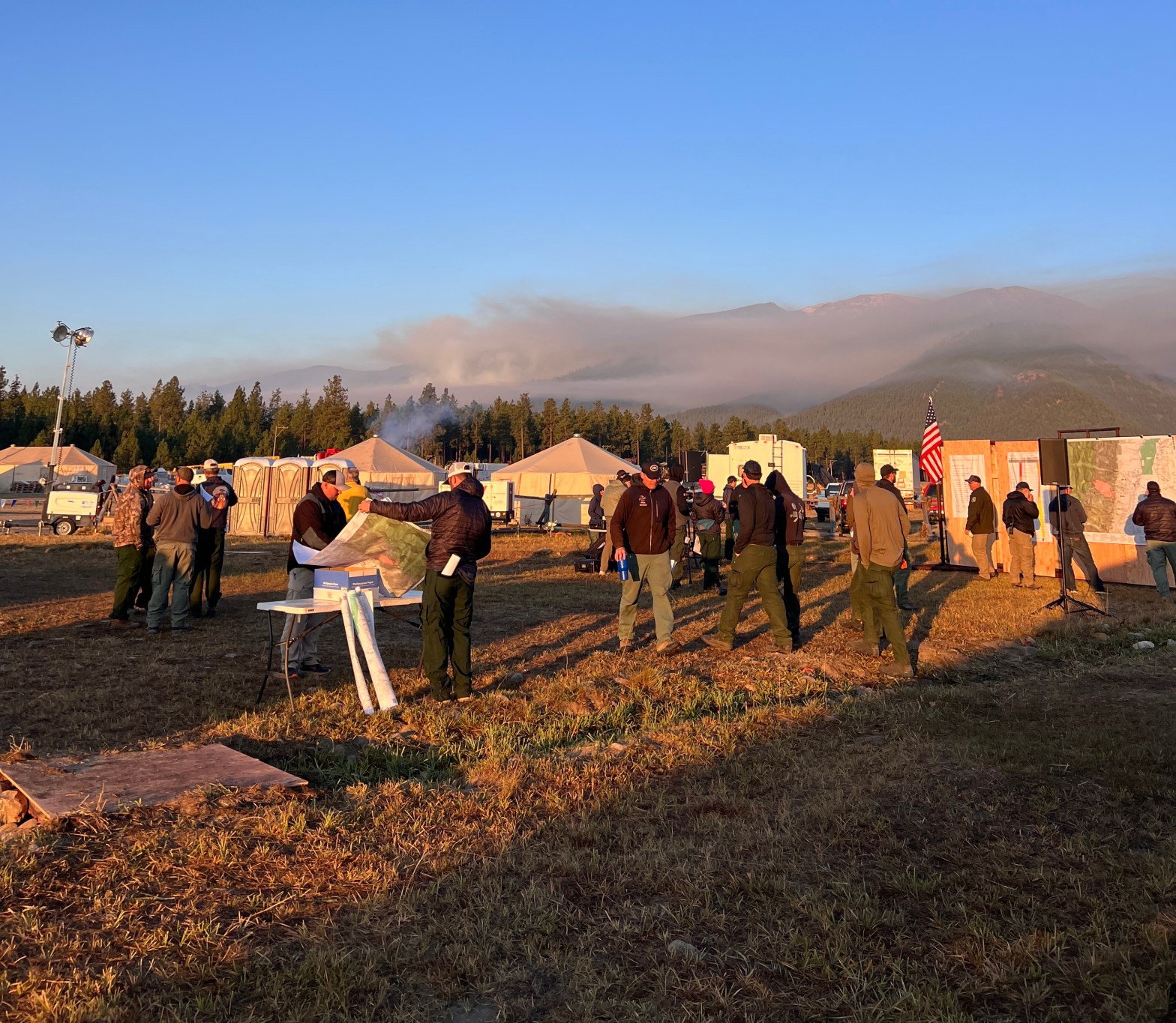 People walk around an incident command post set up in a field looking at maps. Wildfire smoke is seen in the distance and yurts are set up behind the people.