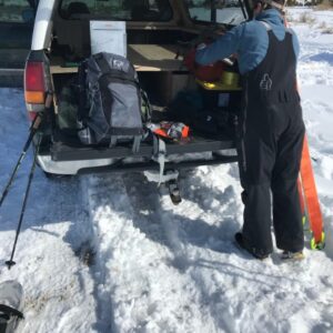 A man in black snowpants getting into the back of a truck. There is snow on the ground and outdoor gear on the tailgate.