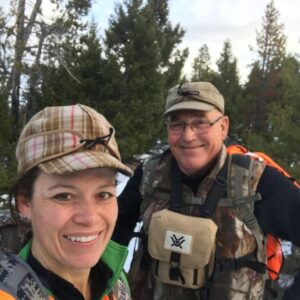 A woman and man with winter hunting gear on smiling in the forest.