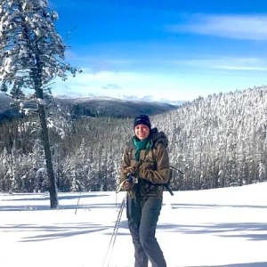 A woman bundled up in winter gear stands on a snowy mountain with pine trees behind her.