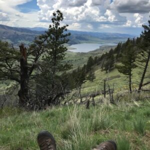 Perspective of someone sitting on a mountain looking out over a valley with a lake in the bottom. Person is  wearing hiking boots are in the bottom of the picture.
