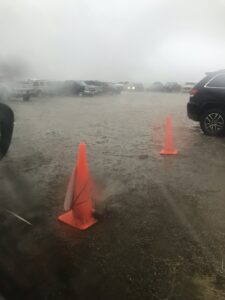 A dirt parking lot at a wildfire incident command post being soaked inundated with rain. Vehicles and orange safety cones are surrounded by water.