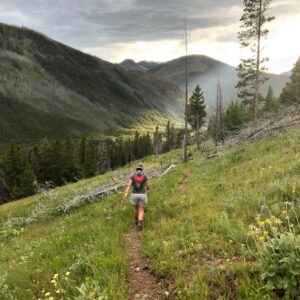 A woman hiking up a trail surrounded by green meadows. Mountains are in front of her.