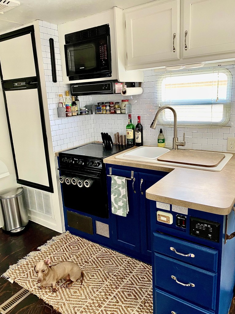 A renovated RV kitchen with a weiner dog sitting on a rug on the floor.