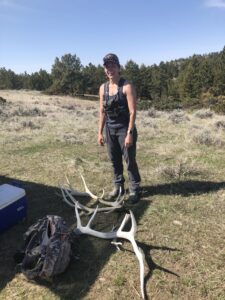 Author standing in front of a pile of elk and deer antlers lying on the ground she found shed hunting. She is wearing a tank top and pants and a set of binoculars in a harness. Sagebrush and trees are in the background.