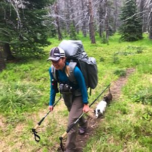 A woman backpacking down a dirt trail holding trekking poles in both hands with full backpack on her back. Two dachshunds, a white and brown one, follow her down the trail. Save weight to have room for your dogs gear.
