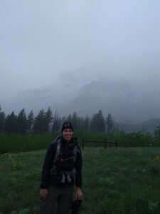 Snowstorm in June in the mountains. Woman wearing warm clothes and a backpack in a field of green with a cloud covered mountain in the background.