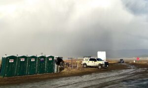 Picture of a row of green porta potties. Next to it is a BLM wildland fire emergency vehicle. A dirt road recently rained on is in front of the truck and porta-potties. A rain storm is occurring in the background.