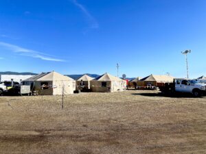 Picture of a bright, blue sky, with 5 tan portable yurts set up. A truck is in the foreground. Brown ground is in the bottom half of the picture.