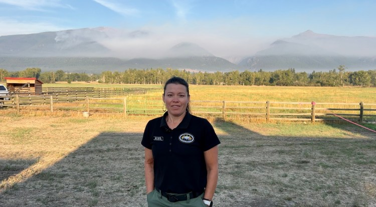 Jeni Garcin stands in a field with wildfire smoke in the distance behind her.