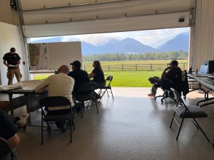 Members of an incident management team strategize inside a building with the door open. Wildfire smoke is seen on the mountain in the distance.